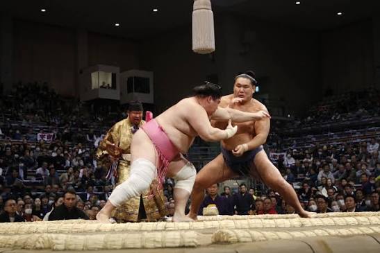Sumo bout in progress on the dohyō with the gyōji referee in gold robes and a full arena watching